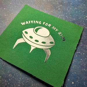 May include: Green fabric patch with silver foil lettering that reads "Waiting for my ride". A silver foil spaceship is in the center of the patch.