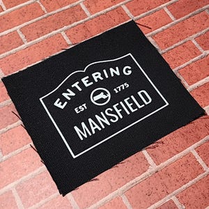 May include: Black fabric patch with white text that reads "Entering Est. 1775 Mansfield" with a circle containing a stylized map of the state of Connecticut.