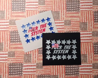 May include: Two rectangular fabric patches. One is off-white with blue stars and red text reading FUCK THE SYSTEM with a small American flag icon. The other is black with silver stars and silver text reading FUCK THE SYSTEM with an American flag icon.