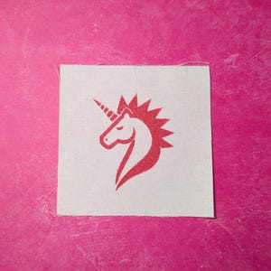 Puede incluir: Un parche de hierro con la silueta de una cabeza de unicornio roja sobre un fondo de tela blanca.