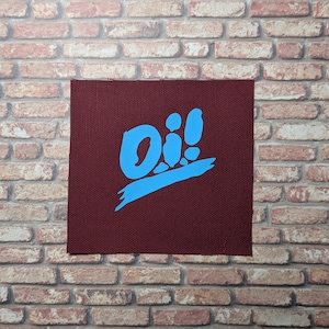 Puede incluir: Un parche de tela marrón con un gráfico azul "Oi!" El gráfico está estilizado con un trazo de pincel azul debajo del texto.