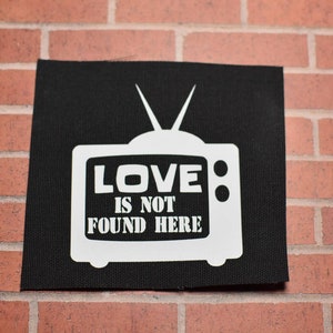 May include: A square black fabric patch featuring a white screen-printed graphic of a vintage television set with two antennas. Inside the television screen, the text reads LOVE IS NOT FOUND HERE in bold white capital letters.