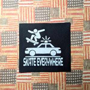 Op de afbeelding: Zwart-witte strijkpatch met een silhouet van een persoon die skateboard op een politieauto. De tekst "Skate Everywhere" is onder de afbeelding gedrukt.