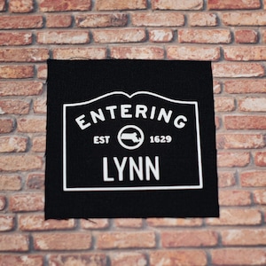 May include: Black fabric patch with white text that reads "Entering Est 1629 Lynn" with a white outline of the state of Massachusetts inside a circle.