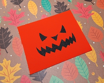 May include: A red fabric patch with a black jack-o'-lantern face. The face has a wide, toothy grin and two small, pointed eyes.