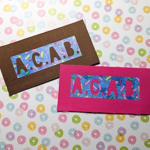 Op de afbeelding: Twee stoffen patches met het acroniem "A.C.A.B." gespeld in witte letters op een blauwe achtergrond met kleurrijke sprinkles. De patches zijn bruin en roze.