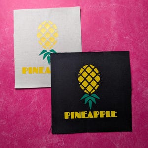 May include: Two fabric patches with a pineapple design. The first patch is white with a gold pineapple and the text "PINEA" in yellow. The second patch is black with a yellow pineapple and the text "PINEAPPLE" in yellow.