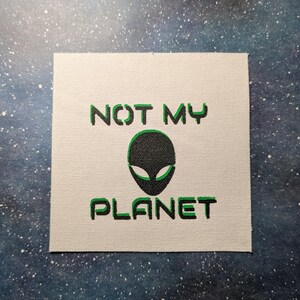 Op de afbeelding: Een witte canvas patch met groene tekst die "NOT MY PLANET" luidt en een zwarte alien-kopgrafiek.