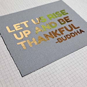Puede incluir: Una tela de lona gris con las palabras "Let us rise up and be thankful -Buddha" en letras doradas, verdes y marrones.