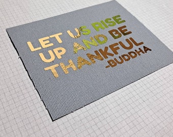 May include: A gray canvas fabric with the words "Let us rise up and be thankful -Buddha" in gold, green, and brown lettering.