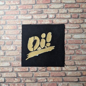May include: A black fabric patch with a gold "Oi!" graphic. The graphic is stylized with a thick, white outline and a gold brushstroke underneath.