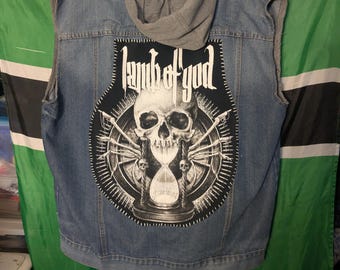 May include: A blue denim sleeveless vest with a grey hood and a large black and white back patch featuring a skull, an hourglass, and the text Lamb of God. The vest has a button-up front, chest pockets, and raw-edged armholes, showcasing a classic denim jacket construction.