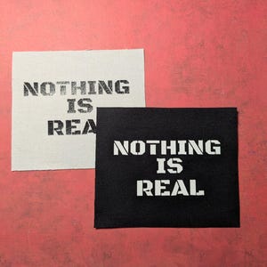 Puede incluir: Dos parches de tela con el texto "Nothing is Real" impreso en negro sobre fondo blanco y blanco sobre fondo negro.