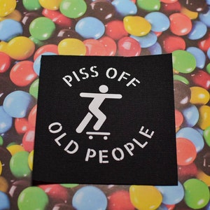 Może przedstawiać: Czarna łatka z białym tekstem, który brzmi "Piss Off Old People" z postacią patyczaka jeżdżącego na deskorolce w środku.