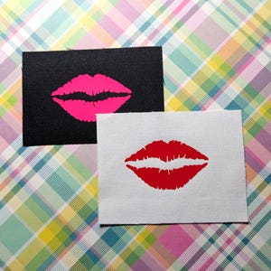 Può includere: Due toppe di tessuto con un design a stampa labbra. Una toppa è nera con una stampa labbra rosa. L'altra toppa è bianca con una stampa labbra rossa.