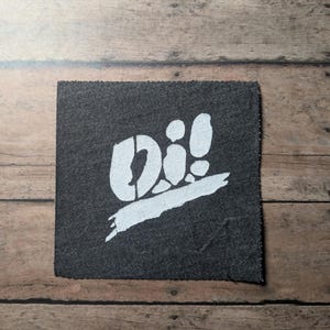 May include: A square-shaped dark gray fabric patch featuring a white screen-printed graphic. The design displays the text "Oi!" in a bold, stylized font positioned above a thick, horizontal brushstroke element that spans across the lower section.