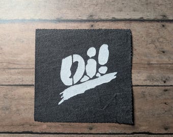 May include: A square-shaped dark grey fabric patch featuring a white screen-printed graphic. The design displays the text "Oi!" in a bold, stylised font positioned above a thick, horizontal brushstroke element that spans across the lower section.