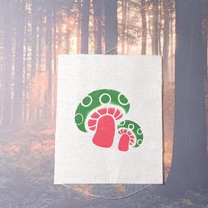May include: A white fabric patch with a green and red mushroom design. The mushroom has a green cap with white circles and a red stem.