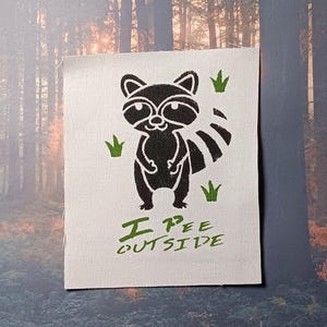 Puede incluir: Un parche de tela blanco con una silueta negra de un mapache con el texto "I pee outside" en verde.