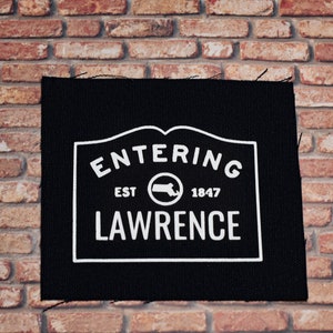 May include: Black fabric patch with white text that reads "Entering Est 1847 Lawrence" with a white outline of the state of Massachusetts inside a circle.