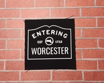 May include: Black and white patch with the text "Entering Est. 1722 Worcester" on a white sign with a black outline. The sign has a rounded top and a small black silhouette of the state of Massachusetts.