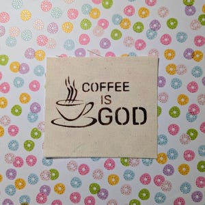 Op de afbeelding: Een witte stoffen patch met bruine tekst die "COFFEE IS GOD" zegt en een bruine illustratie van een koffiekopje met stoom die eruit opstijgt.