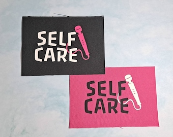 May include: Two rectangular patches, one black and one pink, each with the words "SELF CARE" in white block letters. A pink and white vibrator graphic is on each patch. The patches are on a light blue background.
