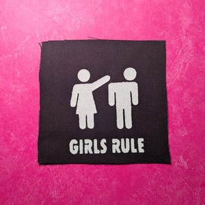 Puede incluir: Un parche de tela negro con figuras de palo blancas de una mujer señalando a un hombre. El texto "GIRLS RULE" está impreso debajo de las figuras.