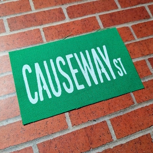 May include: A green fabric patch with white text that reads "Causeway St."