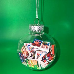 Könnte beinhalten: Transparenter Weihnachtsbaumschmuck aus Glas, gefüllt mit Miniatur-Filmcovern. Der Schmuck hängt an einer silbernen Kette vor einem grünen Hintergrund. Die Filmcover sind farbenfroh und zeigen verschiedene Titel.