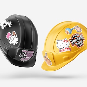 Pode incluir: Dois capacetes de constru&ccedil;&atilde;o, um preto e outro amarelo, decorados com v&aacute;rios autocolantes. O capacete preto tem autocolantes de gestos com as m&atilde;os. O capacete amarelo tem um autocolante Hello Kitty e o texto "Let's Keep the Dumbfuckery to a Minimum Today."