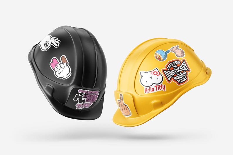 Pode incluir: Dois capacetes de constru&ccedil;&atilde;o, um preto e outro amarelo, decorados com v&aacute;rios autocolantes. O capacete preto tem autocolantes de gestos com as m&atilde;os. O capacete amarelo tem um autocolante Hello Kitty e o texto "Let's Keep the Dumbfuckery to a Minimum Today."