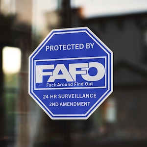 以下が含まれることがあります： 「PROTECTED BY FAFO Fuck Around Find Out」と「24 HR SURVEILLANCE 2ND AMENDMENT」のテキストが入った、青と白の八角形の標識。ガラス面に貼られています。