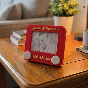 Puede incluir: Un marco rojo estilo Etch-A-Sketch con los nombres "Justin & Isabella" en letras doradas. El marco contiene un boceto a lápiz de una pareja besándose. Las palabras "Etch A Sketch" están impresas en la parte inferior.
