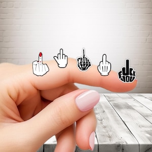 May include: Five middle finger stickers in different designs. One has a red painted fingernail, another is a skeleton hand, and the others are outlined in black. The stickers are on a finger.