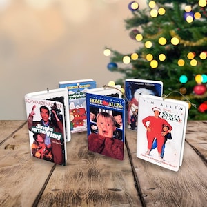 Könnte beinhalten: Eine Sammlung von Weihnachtsbaumschmuck in Form von Filmcovern. Die Ornamente zeigen Filmplakate von Weihnachtsfilmen wie "Jingle All the Way", "Home Alone" und "The Santa Clause". Sie haben weiße Ränder und eine goldene Aufhängeschnur.