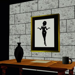 May include: A framed black and white silhouette of a woman wearing a necklace and a skirt, standing with her arms outstretched. The frame is gold and the background is a brick wall. A black vase and a brown mug are on a wooden table in the foreground.