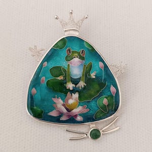 May include: A silver triangular pendant with a green frog sitting on a lily pad, surrounded by pink flowers and green leaves. The pendant has a crown on top and a green gemstone at the bottom.