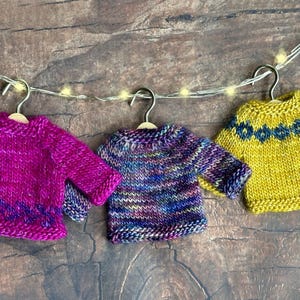 Three colorful miniature hand knit sweater ornaments lined up on a wood background. Each mini sweater is a different color and has a different pattern.