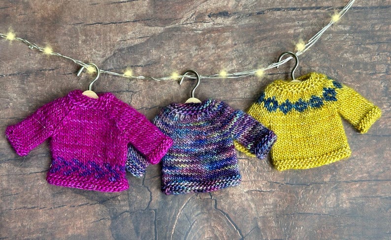 Three colorful miniature hand knit sweater ornaments lined up on a wood background. Each mini sweater is a different color and has a different pattern.