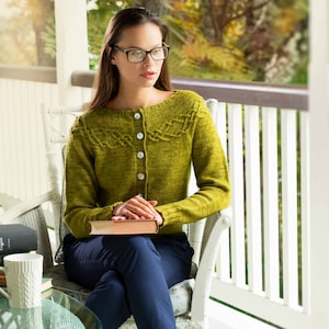May include: A woman wearing a green knitted cardigan with a cable knit pattern sits on a white wicker chair holding a book. She is wearing blue jeans and has glasses on.