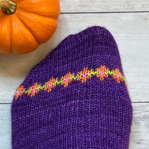 Puede incluir: Primer plano de un calcet&iacute;n morado tejido con una banda decorativa de diamantes naranjas, rosas y verdes. Una calabaza naranja se encuentra en la esquina superior izquierda. El calcet&iacute;n est&aacute; hecho de hilo texturizado.