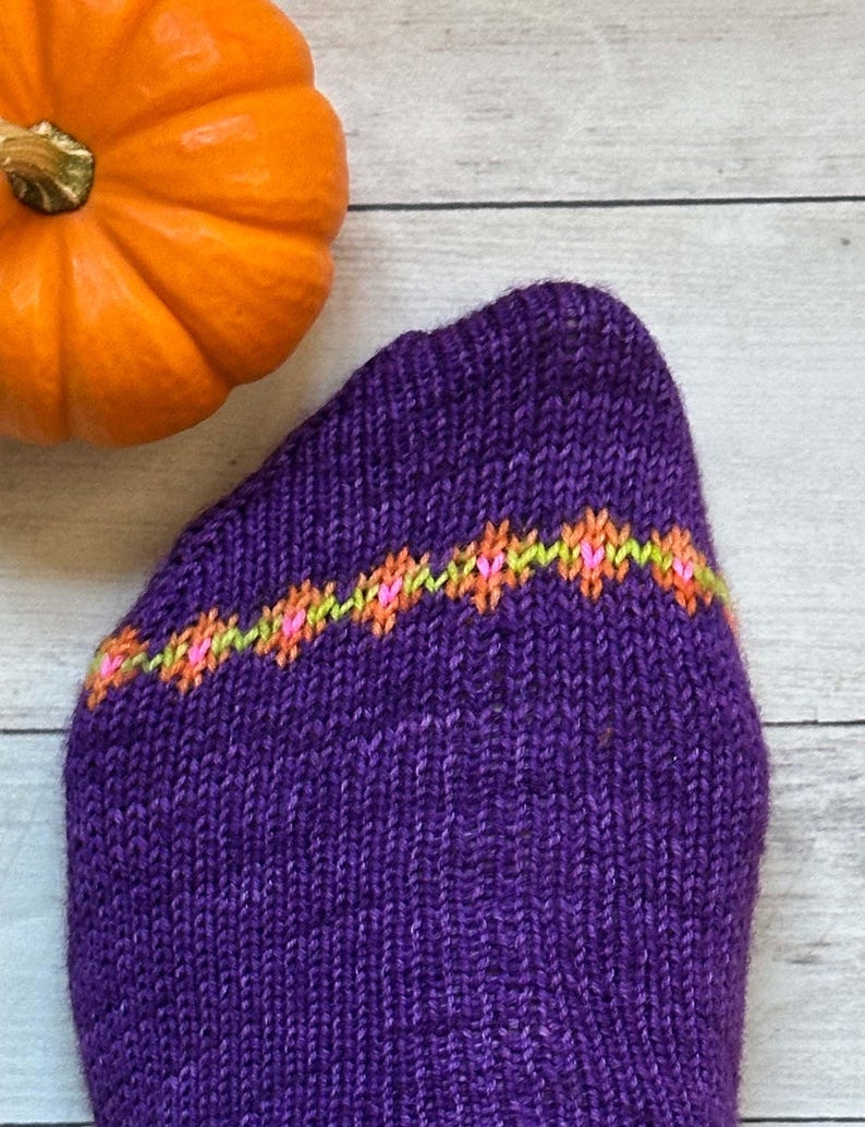 Puede incluir: Primer plano de un calcet&iacute;n morado tejido con una banda decorativa de diamantes naranjas, rosas y verdes. Una calabaza naranja se encuentra en la esquina superior izquierda. El calcet&iacute;n est&aacute; hecho de hilo texturizado.