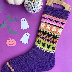 May include: A purple knitted sock with a Halloween pattern featuring bats, ghosts, and jack-o'-lanterns. The sock is displayed on a wooden hanger against a purple background, with decorative cutouts of bats, ghosts, and a pumpkin.