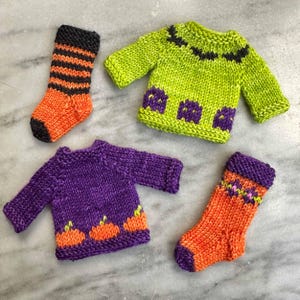 May include: A collection of handmade knitwear items, including two miniature sweaters and two socks. The sweaters are green and purple, with decorative elements. The socks are orange with black and purple accents. The items are likely for a doll or small toy.