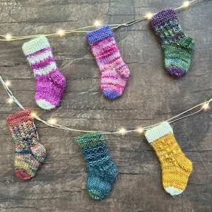 May include: Six colorful knitted stockings with different patterns hang from a string of lights. The stockings are made of yarn in shades of pink, purple, green, yellow, blue, and red.