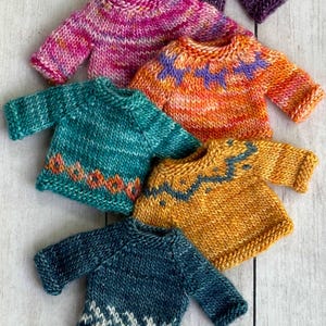 Könnte beinhalten: Eine Sammlung von handgestrickten Miniaturpullovern in verschiedenen Farben. Die Pullover weisen Muster und Designs in Rosa-, Orange-, Türkis-, Gold- und Blautönen auf. Die Pullover sind auf einer hellen Holzoberfläche angeordnet.