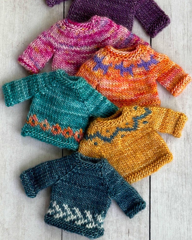 Six colorful miniature hand knit sweater ornaments on a white wood background. Each mini sweater is a different color and has a different pattern.