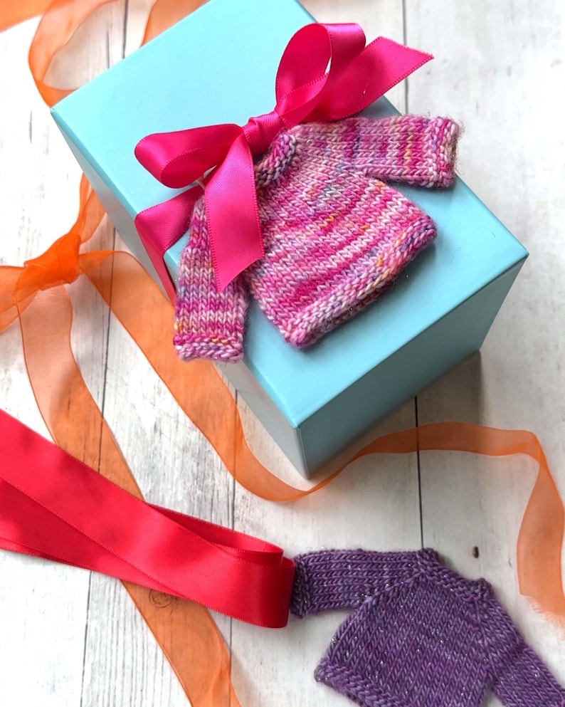 A pink miniature hand knit sweater adorning a gift with a bright pink bow.