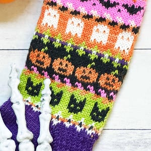 Puede incluir: Un calcet&iacute;n de Halloween tejido de color p&uacute;rpura y multicolor con murci&eacute;lagos, fantasmas, calabazas y gatos. El calcet&iacute;n presenta un pu&ntilde;o acanalado y un dise&ntilde;o estampado en rosa, naranja, negro y verde. Una mano de esqueleto decorativa est&aacute; en primer plano.
